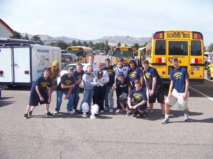 Meridian drumline students holding a competition trophy in front of school buses